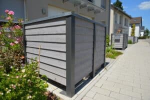 Mülltonnenverkleidung aus Holz und Metall vor Mehrfamilienhaus in moderner Siedlung mit Blumenbeet