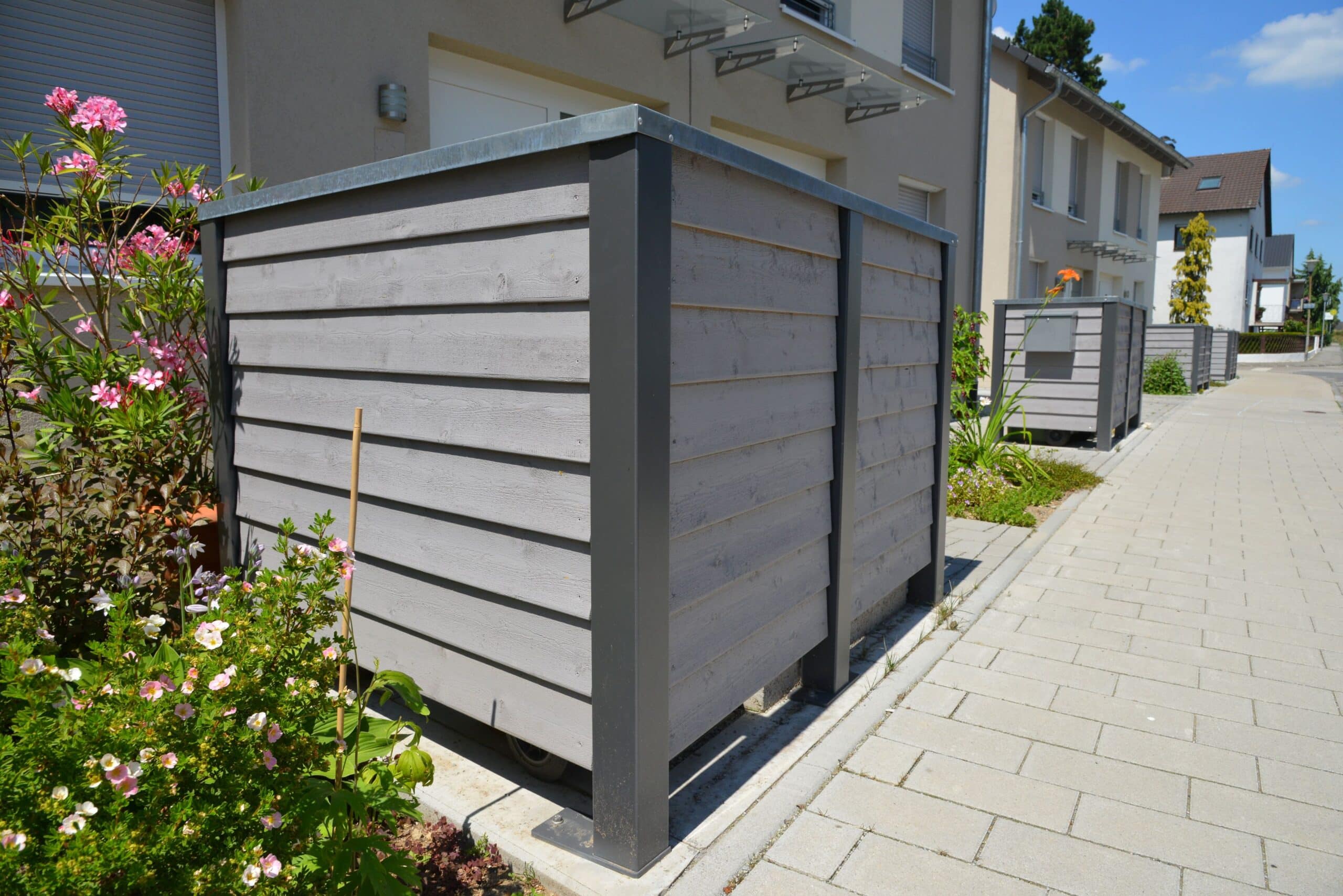 Mülltonnenverkleidung aus Holz und Metall vor Mehrfamilienhaus in moderner Siedlung mit Blumenbeet